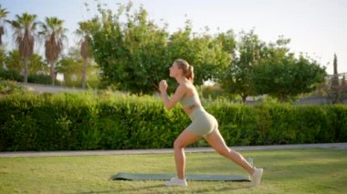 Genç ve atletik bir kadın açık havada renkli bir yoga minderi üzerinde egzersiz yapıyor. Etkileyici fitness rutinini sergilerken doğa tarafından çevrelenmiş sıcak ve güneşli günün tadını çıkarıyor.