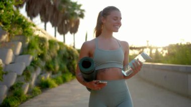 Moda kıyafetleri içindeki genç bir kadın, doğa tarafından çevrelenmiş, açık hava ve sağlık koşullarına olan bağlılığını gösteren parlak bir yoga minderi ve gösterişli bir su şişesi kullanırken kendinden emin bir şekilde gülümsüyor.