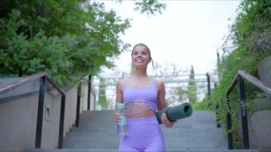 Zinde ve kendinden emin bir kadın yoga minderini ve ferahlatıcı bir içeceği coşkuyla taşır ve çevresini çevreleyen canlı yeşilliğe karşı aktif ve sağlıklı yaşam tarzını mükemmel bir şekilde sergiler.