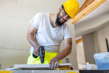 Güvenlik kaskı ve parlak yelek giyen dinamik bir erkek inşaat işçisi, yoğun bir inşaat alanındaki bir ev yenileme projesinde ustaca bir matkap kullanarak uzmanlığını sergiliyor.