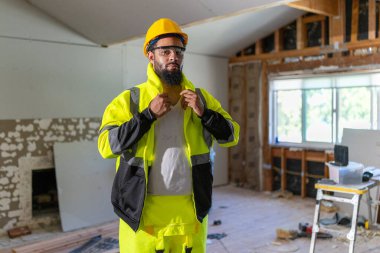 Parlak güvenlik yeleği ve miğferi olan neşeli bir inşaat işçisi, tadilat yapılan bir evde güvenle duruyor. Alet ve malzemeler arasında odaklanma ve profesyonelliğini gösteriyor.