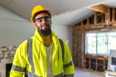 Emniyet yeleği ve miğfer giyen neşeli bir adam tadilat alanında kendine güveni tam bir şekilde duruyor, inşaat ve işçilikle ilgili coşkulu ruhu ve kendini adamışlığı somutlaştırıyor.
