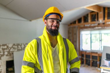 Parlak güvenlik yeleği ve sağlam bir miğfer giyen neşeli bir inşaat işçisi, sürmekte olan projeye kendini adamış ve uzmanlığını sergileyerek, tadilattan sorumlu bir inşaat sahasında güven içinde duruyor.