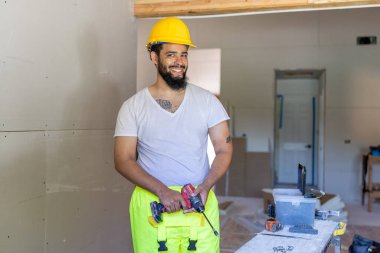 Güvenlik kaskı ve yansıtıcı yelek giymiş neşeli genç bir inşaat işçisi evi yenilerken gülümseyerek profesyonellik, adanmışlık ve marifet gösteriyor.