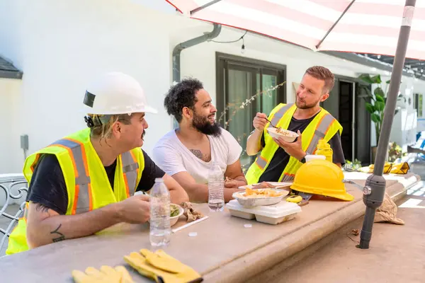 Bir projede güneşli bir öğle yemeği molası sırasında, çeşitli inşaat işçileri ve bir mimar kahkahaları paylaşır, yemekten zevk alır ve canlı sohbetler yapar, işbirliği ve arkadaşlığı teşvik eder.
