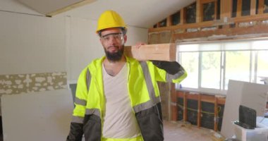 Canlı bir güvenlik yeleği ve miğferi olan neşeli, genç bir inşaat işçisi gururla ahşap bir kalas tutuyor. Binaların görünümünü ve işlevselliğini geliştirmek için tadilat projesine girişiyor.