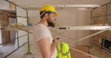 Parlak bir güvenlik yeleği ve koruyucu bir kask takan erkek inşaat işçisi, yenilenmiş bir alanda güvenle duruyor. Telefon, çeşitli görevler ve kalite sonuçlarına odaklanıyor. 