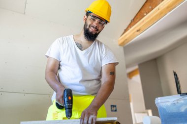 Sarı yelek ve kask giyen neşeli bir inşaat işçisi hareketli bir ortamda işleri ustalıkla idare ediyor. Profesyonelliğini ve etkileyici işçiliğini her detayıyla gösteriyor.