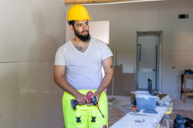 Güvenli bir kask ve parlak giysiler giyen genç bir erkek inşaat işçisi, bir ev yenileme projesinin ortasında duruyor, sanatında kendini adamış ve profesyonelliğini sergiliyor.