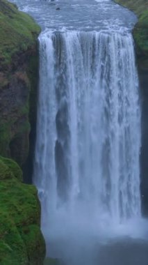 Dramatik hava görüntüsü Ünlü Skogafoss şelalesinin güçlü akışını, bulutlu bir gökyüzünün altında, doğanın saf güzelliğiyle çevrili yemyeşil bir uçurumdan aşağı yuvarlanışını yakalar. İzlanda, Skogar 