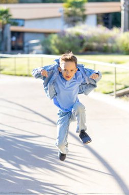 Açık mavi kıyafetli neşeli genç bir çocuk dışarıda güneşin altında koşup oynuyor. Bulaşıcı coşkusu ve mutluluğuyla herkesi neşelendiren parlak bir gülümseme paylaşıyor.