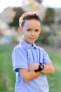 Canlı bir açık hava ortamında, genç bir çocuk kollarını kavuşturmuş, şık mavi bir polo ve parlak siyah bir saat takıyor, neşeli mizacını yansıtan büyüleyici bir gülümseme sergiliyor.