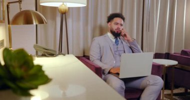 Sivri bir takım elbise giymiş şık, genç Afrikalı Amerikalı adam, modern, çağdaş ofis ortamında dizüstü bilgisayarı üzerinde ustalıkla çalışırken aktif olarak sanal bir toplantı yapıyor.