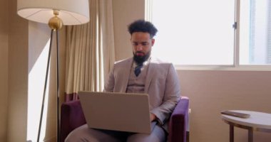 Zarif, açık renkli bir takım elbise giymiş Afro-Amerikalı bir adam, doğal ışıkla güzelce aydınlatılmış sofistike bir otel odasında dizüstü bilgisayarına derinden odaklanmıştır. Takım elbiseli iş adamları