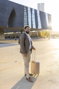 Entelektüel profesyonel giysiler içinde Afrika kökenli Amerikalı bir işadamı, gün batımında geniş bir şehir manzarasında dikilirken kendine güvenen bir çanta tutar.