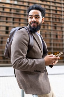 Afro-Amerikalı bir işadamı telefonuna bakarken gülümsüyor, şık kahverengi bir ceket giyiyor ve şık bir sırt çantası taşıyor, hareketli bir şehir ortamında kendine güven ve profesyonellik yayıyor.