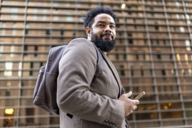 Kendine güvenen, iyi giyimli bir Afro-Amerikan işadamı kalabalık bir şehir ortamında elinde akıllı bir telefonla duruyor ve şık bir sırt çantası taşıyor, kararlılığı ve kariyer başarısını yansıtıyor.