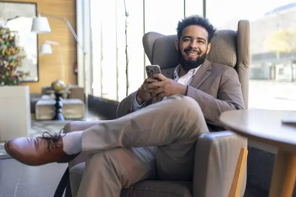 Afrikalı Amerikalı bir işadamı şık bir sandalyeye oturarak akıllı telefonunu kullanır. Sağlıklı bir iş hayatı dengesini vurgulayan rahat ama profesyonel bir yaşam tarzı sergiler.