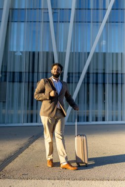 İyi giyimli, dengeli bir Afro-Amerikan işadamı, modern bir mimari yapının önünde elinde bir bavulla, seyahat etmeye ve profesyonel görünmeye hazır bir şekilde duruyor.