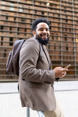 Moda ve şık bir kıyafet giymiş neşeli bir Afro-Amerikan işadamı, hareketli bir şehir ortamında, iyimserlik ve kararlılık yayarak akıllı telefonunu kendine güvenle tutuyor.
