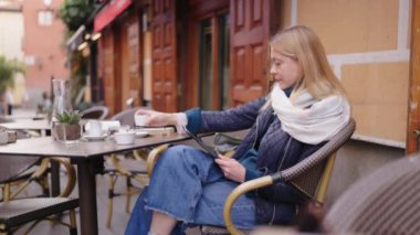 Genç bir kadın açık bir kafede oturuyor, tablet kullanırken kahvesini yudumluyor, büyüleyici bir sokak atmosferiyle çevrili.