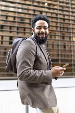 Kendine güvenen, güler yüzlü, kahverengi takım elbiseli bir Afro-Amerikan işadamı elinde bir akıllı telefon tutuyor. Parlak, modern bir şehir zeminine karşı pozitiflik ve profesyonellik yayıyor.