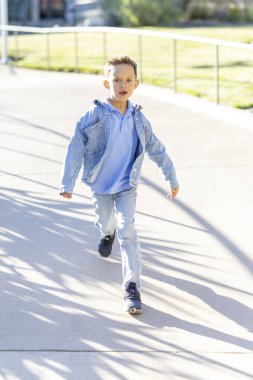 Açık mavi bir elbise ile tamamlanmış şık bir kot ceket giymiş genç bir çocuk, güzel, güneşli bir günde neşeli bir şekilde parkta koşar, havadaki heyecan ve kahkahalarla