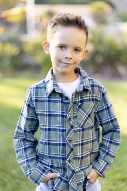 Çekici genç bir çocuk renkli bir ekose gömlekle dışarı poz verirken güneşli park arka planı onun kaygısız davranışını büyük ölçüde artırıyor ve etrafında neşeli bir atmosfer yaratıyor.