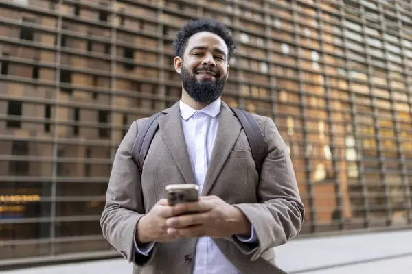 Gülümseyen bir Afro-Amerikan işadamı, elinde telefonla, profesyonelliği ve kendine güveni yansıtan çarpıcı modern mimariye karşı güvenle dışarıda duruyor.