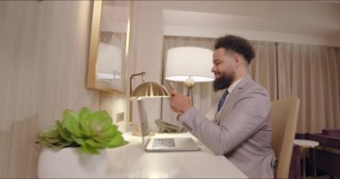 İyi giyimli bir adam, modern bir otel odasına kurulmuş şık bir şekilde tasarlanmış bir masada telefonuyla uğraşırken, canlı ve üretken bir atmosferi etkili bir şekilde sergileyerek güvenle gülümsüyor.