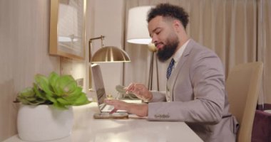 Zarif takım elbiseli başarılı bir işadamı, modern masasında oturur. Dizüstü bilgisayarıyla uğraşırken gülümser ve kariyerinin başarılarını ve kişisel motivasyonunu yansıtır.