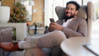 Gülümseyen Afrikalı Amerikalı profesyonel, modern bir salonda rahat bir şekilde oturmakta, akıllı telefonuyla aktif bir şekilde meşgul olurken, atmosferi geliştiren şık bir dekorla çevrelenmiştir.