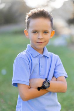Çekici genç bir çocuk, açık hava ortamında kollarını kavuşturmuş açık mavi polo tişört giymiş, masum ve neşe saçan neşeli kişiliğini sergiliyor.
