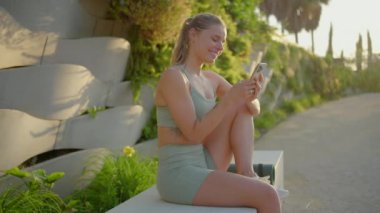 Neşeli bir fitness kadını telefonunu kullanırken keyifli bir an yaşar. Etrafı huzurlu ve güzel bir çevreyle çevriliyken aktif ve sağlıklı bir yaşam tarzını gerçekten somutlaştırıyor.