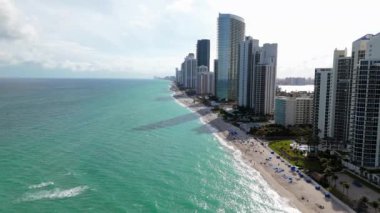 Sunny Isles Sahili 'nin havadan görünüşü savurgan evleri, çarpıcı havuzları ve sallanan palmiye ağaçları olan abartılı bir yaşam tarzını gözler önüne seriyor. Hepsi de görünüşte mükemmel bir günde sıcak güneş ışığıyla aydınlatılıyor. Miami, Florida