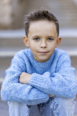Genç bir çocuk düşünceli bir şekilde açık hava ortamında oturuyor, yumuşak mavi bir süveter giyiyor, bu onun çekici ve masum tavrını geliştiriyor, etrafında huzur ve mutluluk hissi uyandırıyor.