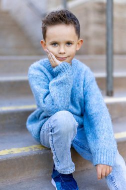 Genç bir çocuk, hoş bir şehir binasının merdivenlerinde, rahat mavi bir süveter ve kot pantolon giyerek, çocukluk sevincini yansıtan, merak ve masumiyet duygusunu arttıran, düşünceli bir şekilde oturur.
