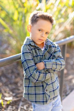 Tarz sahibi genç bir çocuk, gençlik cazibesi ve neşesini somutlaştıran ekose gömleğini ve neşeli ifadesini sergileyen gür yeşilliğin canlı bir zeminine karşı kollarını kavuşturarak güven içinde poz verir.