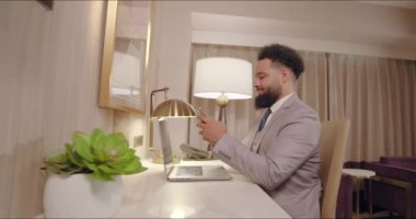 Zarif dekoru olan modern bir ofiste, özel dikim takım elbiseli genç bir profesyonel organize bir masada oturuyor, akıllı telefonlarındaki görevlere odaklanıyor, güçlü bir konsantrasyon ve kararlılık gösteriyor.