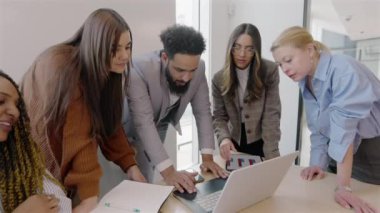 Diverse group of professionals is gathered around a laptop, engaging in a spirited discussion of innovative ideas in a modern office that fosters collaboration, teamwork, and creativity