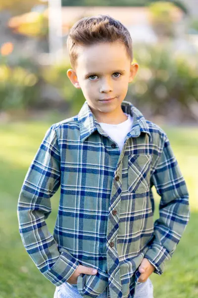 Neşeli ve neşeli bir çocuk şık bir ekoseli gömlek giyerek iki elini de cebine sokup rahat bir şekilde dışarıda duruyor. Doğanın tadını çıkarırken neşeli ve kaygısız kişiliğini sergiliyor.