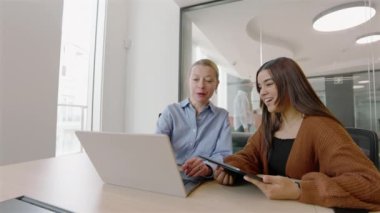 İki meslektaş modern ofis ortamında birlikte çalışmaktan mutluluk duyuyorlar. Toplam verimliliklerini ve çalışma verimliliklerini önemli ölçüde artırmak için bir dizüstü bilgisayar ve tablet kullanıyorlar.