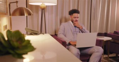 Kendini adamış ve odaklanmış Afro-Amerikan iş adamı düzenli bir şekilde dizüstü bilgisayarı üzerinde çalışıyor, şık, modern bir otel ofisi ortamında, telefonda konuşuyor.