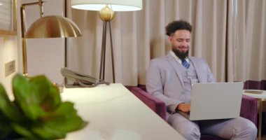 Sofistike ve şık bir ofis ortamında, kendine güveni olan iyi giyimli bir adam modern bir masada oturuyor, dizüstü bilgisayarı üzerinde çalışırken, profesyonellik ve başarı sergiliyor.