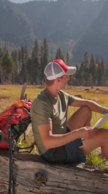 Genç bir adam Yosemite Ulusal Parkı 'nın nefes kesen sakin güzelliğinden zevk alırken dinlenmek için bir dakikasını ayırıyor ve heyecan verici yürüyüş macerası sırasında haritalarını inceliyor.