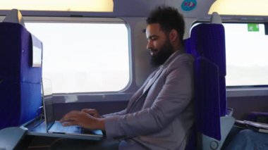 Sivri bir takım elbise giymiş, trende seyahat ederken dizüstü bilgisayarına odaklanmış bir Afro-Amerikan işadamı, günümüzde pek çok kişinin çabaladığı modern iş hayatı dengesini etkin bir şekilde gösteriyor.