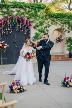 Güzel bir gelin ve şık bir damat neşeli bir şekilde düğün günlerini kutluyorlar. Etrafları canlı çiçeklerle çevrili. Bu da olayları güzelleştiriyor.