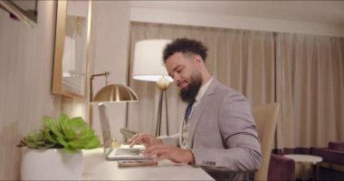 İyi giyimli genç bir adam zarif bir otel odasında dikkatle oturur, dizüstü bilgisayarına dalmış, üretkenliğin özünü somutlaştırır, ve modern, sofistike bir yaşam tarzını özendirir.