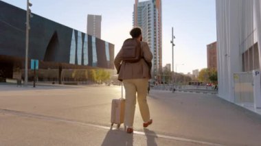 Kararlı iş yolcusu hareket halinde, ustaca bir bavul taşıyor, iş çantası taşıyor, ve önlerine çıkabilecek yeni fırsatlara iyi hazırlanmış, dışarıda sıcak güneş ışığı.