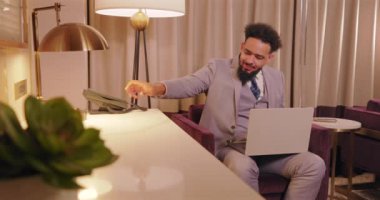 Açık renkli takım elbise giymiş şık bir adam, günümüz modern iş kültürünün özünü mükemmel bir şekilde temsil eden şık bir otel salonunda otururken tamamen dizüstü bilgisayarına odaklanmış durumda.